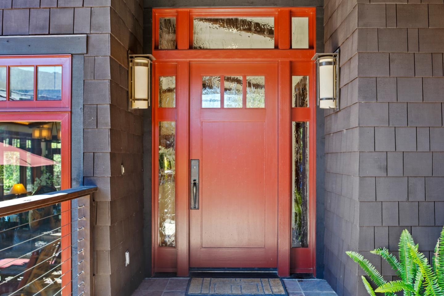 Red front door with glass sidelights and transom, framed by dark shingle siding and porch sconces.