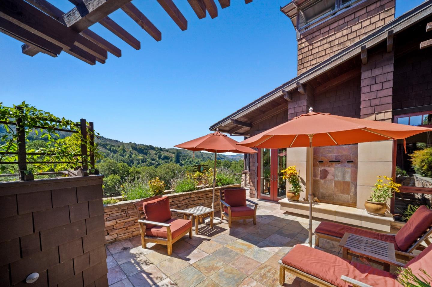 Stone patio with orange umbrellas, red-cushioned chairs, and wooded hills beyond.