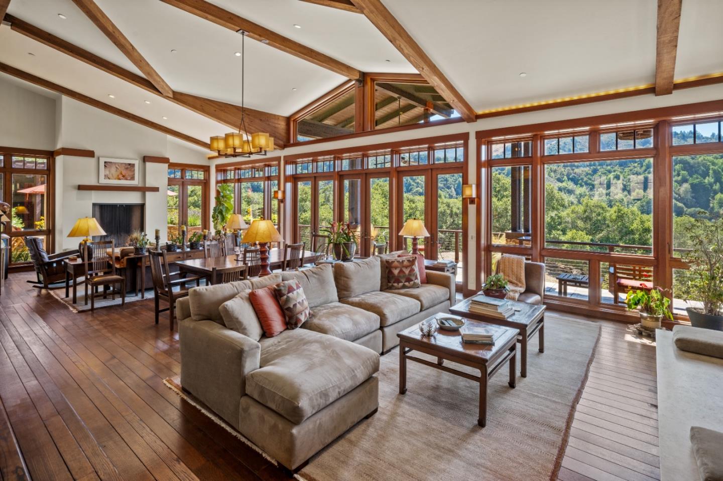 Living and dining room with exposed timber beams, a fireplace, and large windows overlooking wooded hills.