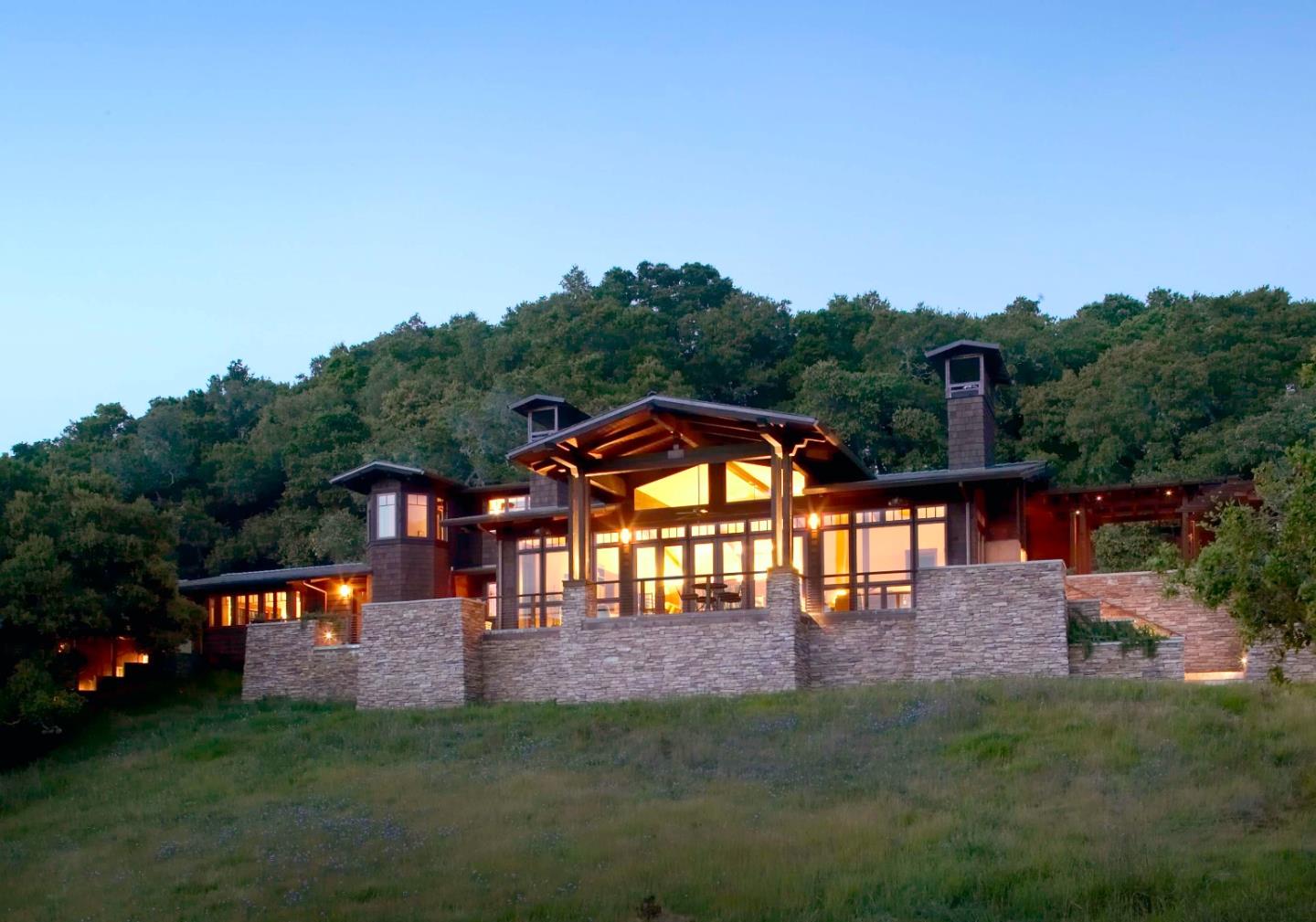 Modern wood-and-stone hillside home glowing at dusk beneath a wooded ridge.