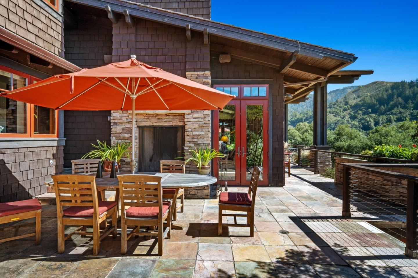 Stone patio with orange umbrella, outdoor dining set, fireplace, and wooded hills beyond.