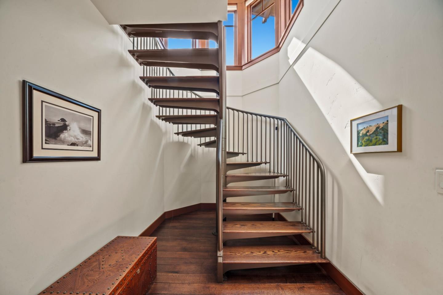 Curved wooden staircase with metal railings beneath tall windows and framed artwork.