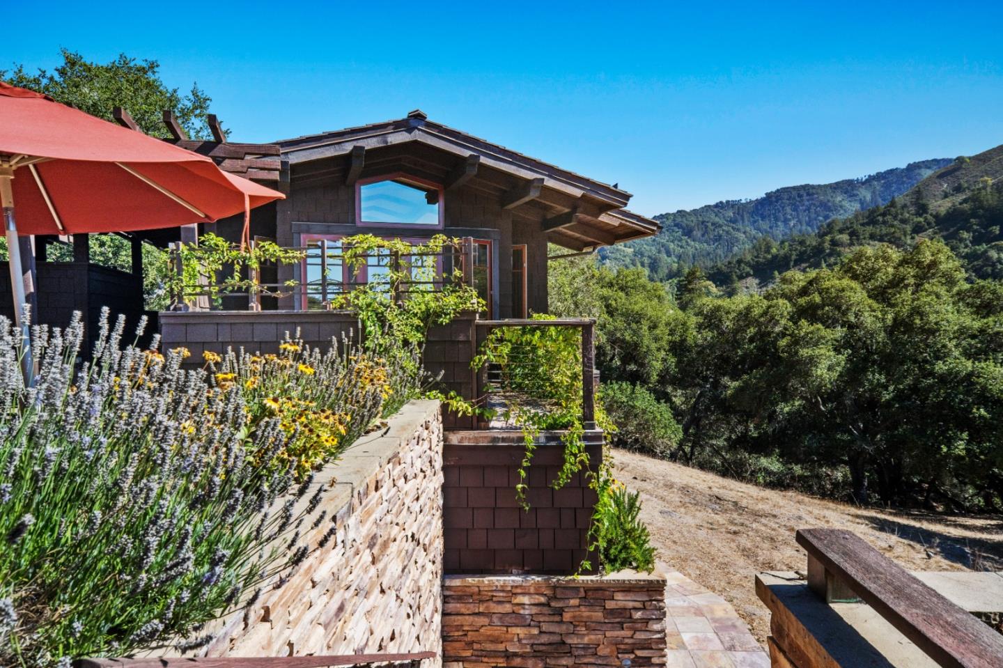 Dark shingled home with a glass-railed deck, red patio umbrella, and wooded hills beyond.