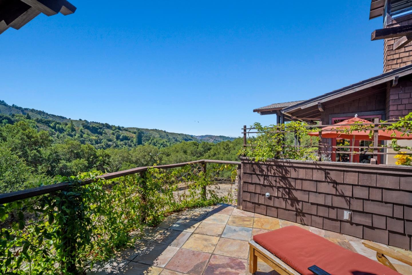Stone-paved deck with a red-cushioned chaise overlooking a wooded valley and rolling hills.