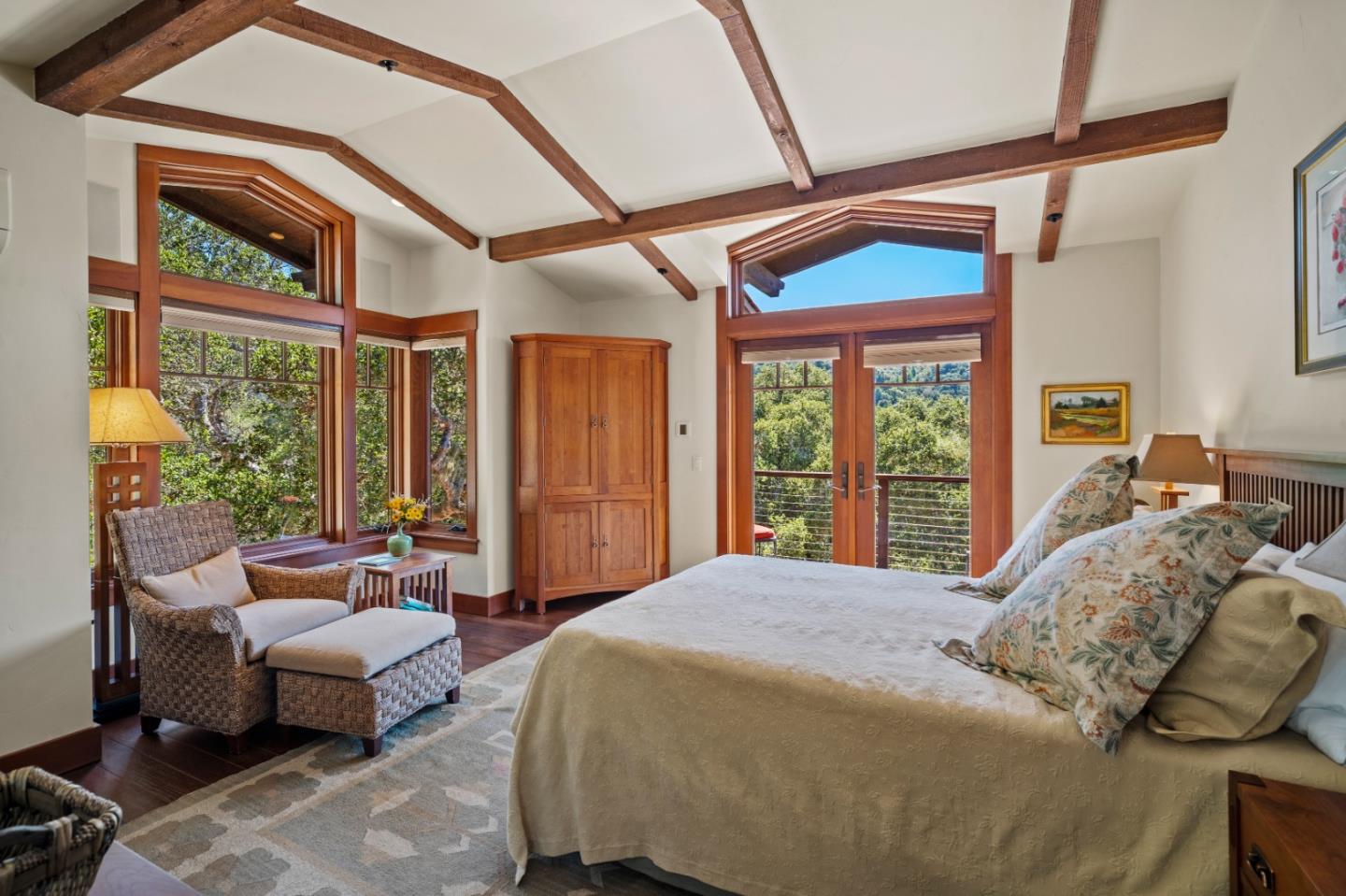 Bedroom with exposed wood beams, large windows, and balcony doors overlooking wooded hills.