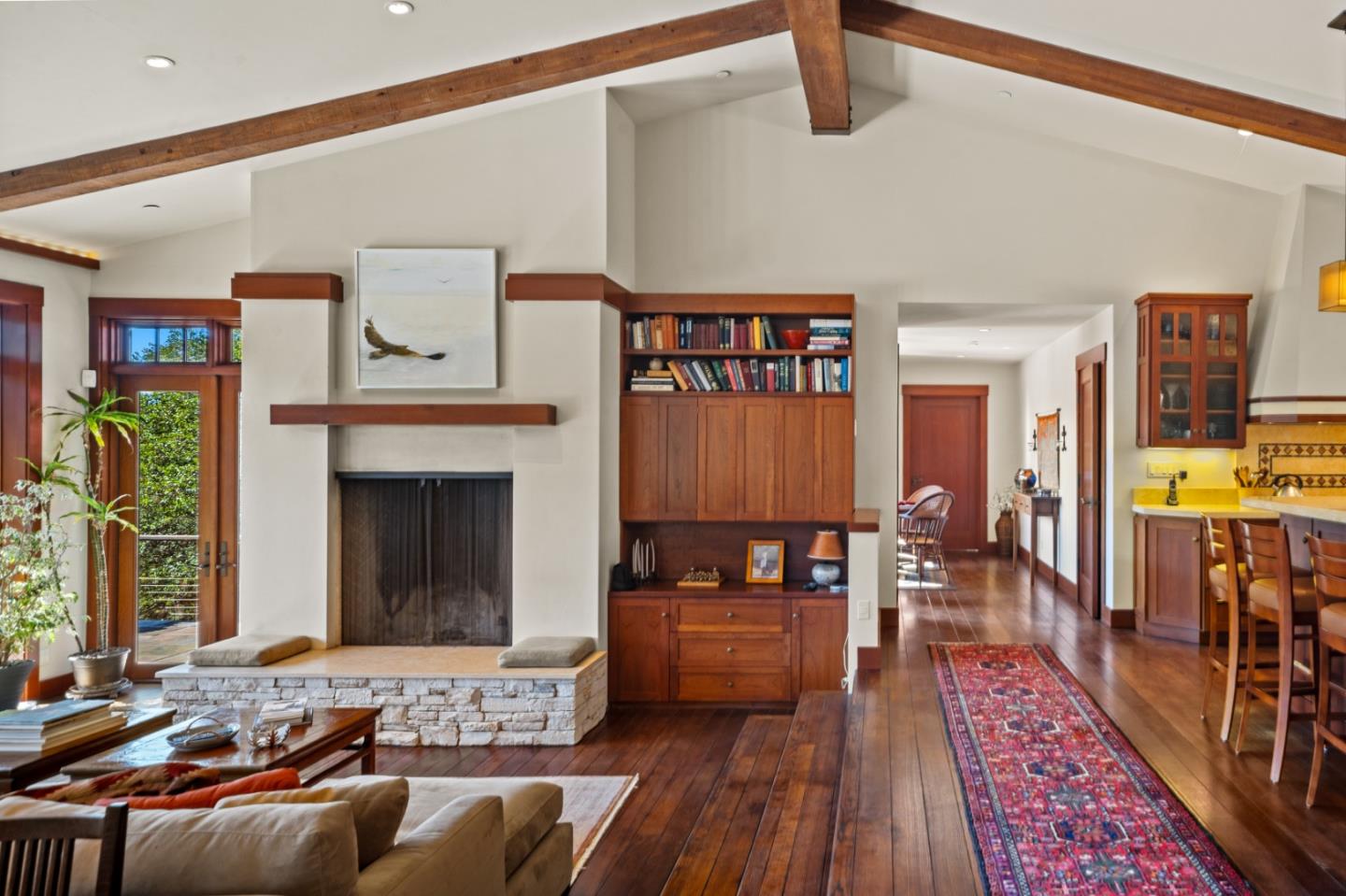 Living room with a stone fireplace, exposed beams, built-in bookshelves, and a long patterned rug.