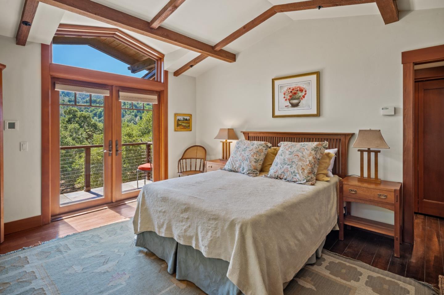 Bedroom with exposed wood beams and glass doors opening onto a balcony overlooking wooded hills.