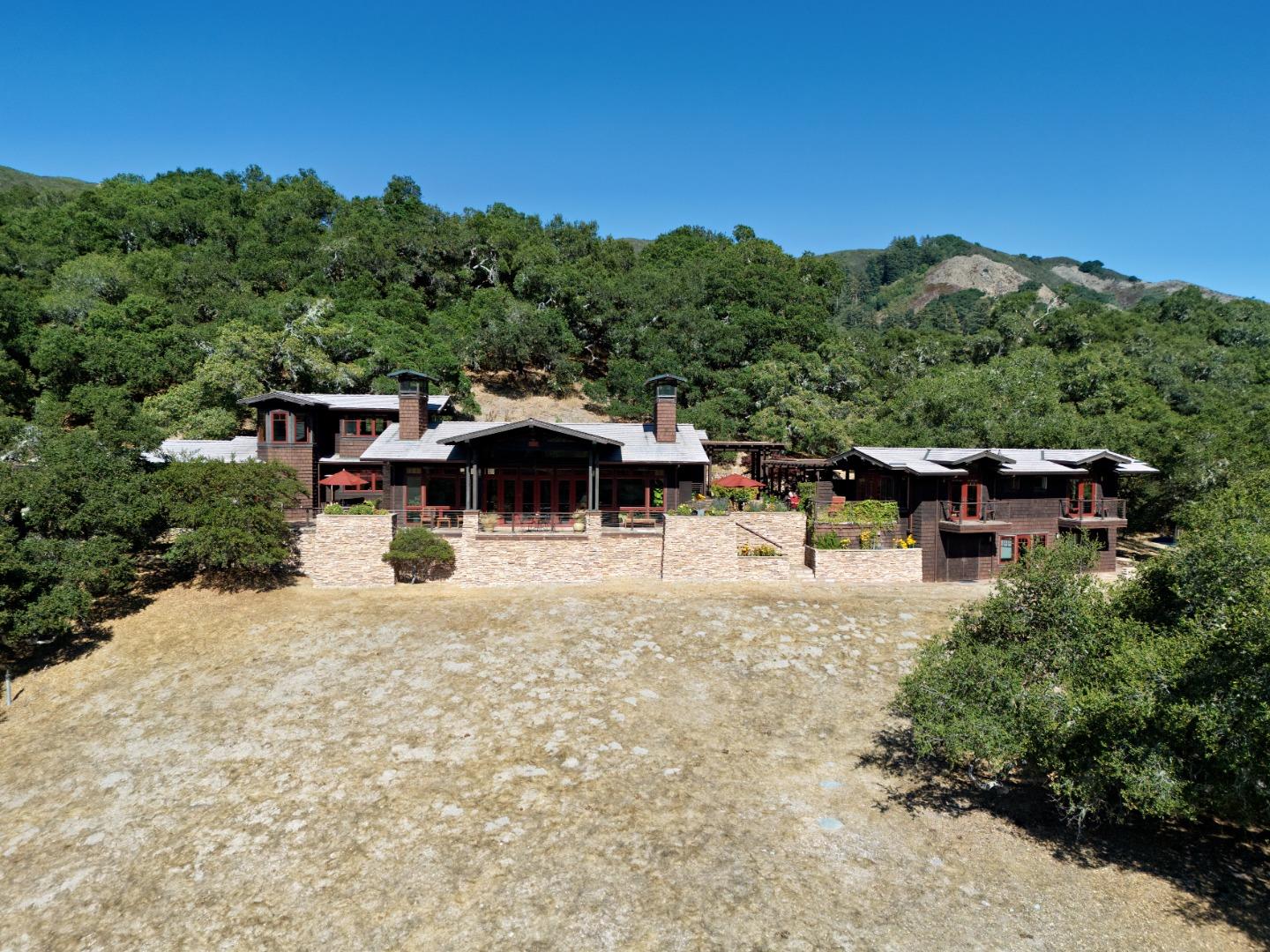 Wood-and-stone home with connected wings and stone garden walls, set against wooded hills.