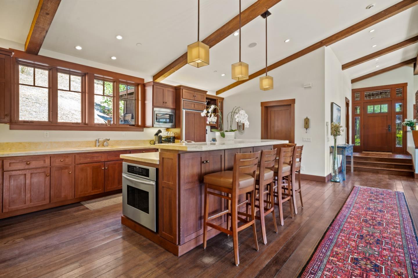 Kitchen with a wood island and bar seating beneath exposed beams and large windows.