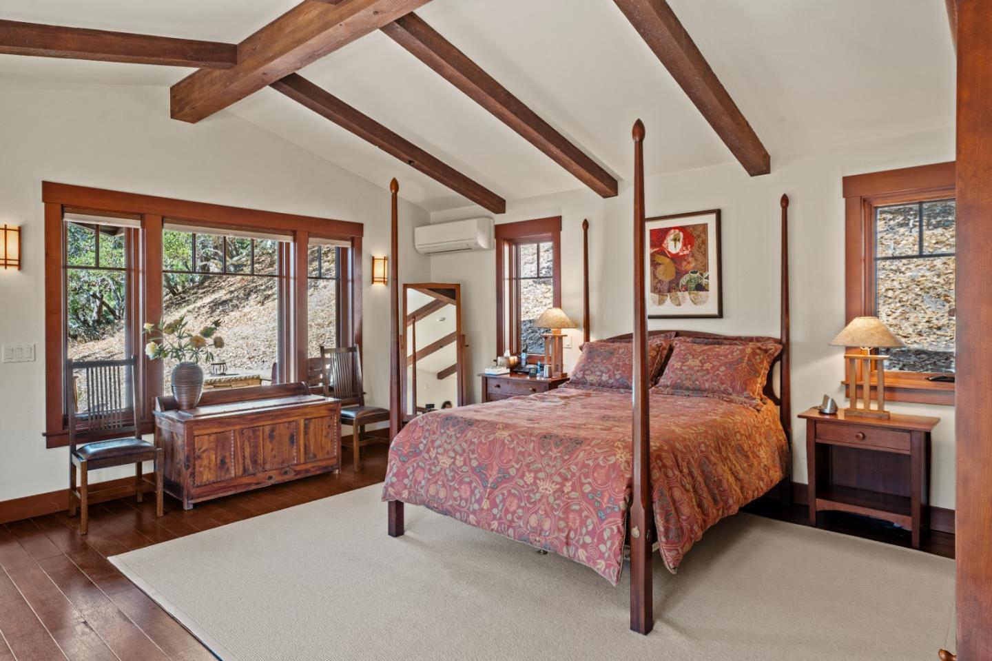 Bedroom with a four-poster bed, exposed wood ceiling beams, and windows overlooking a wooded hillside.