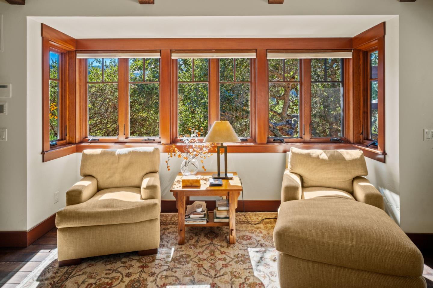 Sitting nook with beige chairs and broad wood-framed windows overlooking leafy greenery.
