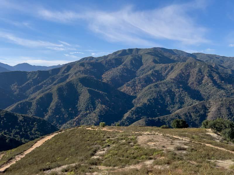 Open hillside with a dirt track overlooking rugged, tree-covered mountains.