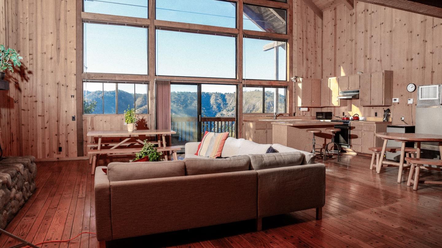Open-plan living room and kitchen with tall windows overlooking wooded mountains.