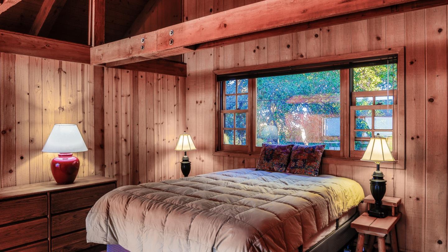 Wood-paneled bedroom with exposed beams, a quilted bed, and a large window overlooking trees.