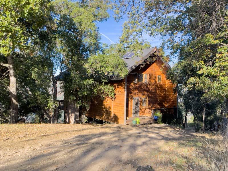 Wood-sided house among mature trees, with rooftop solar panels and a dirt driveway.