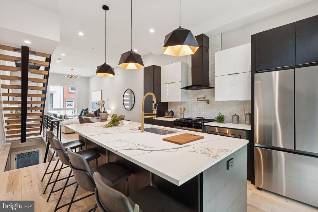 Modern kitchen with a large marble-look island, gold faucet, geometric pendants, and floating staircase.