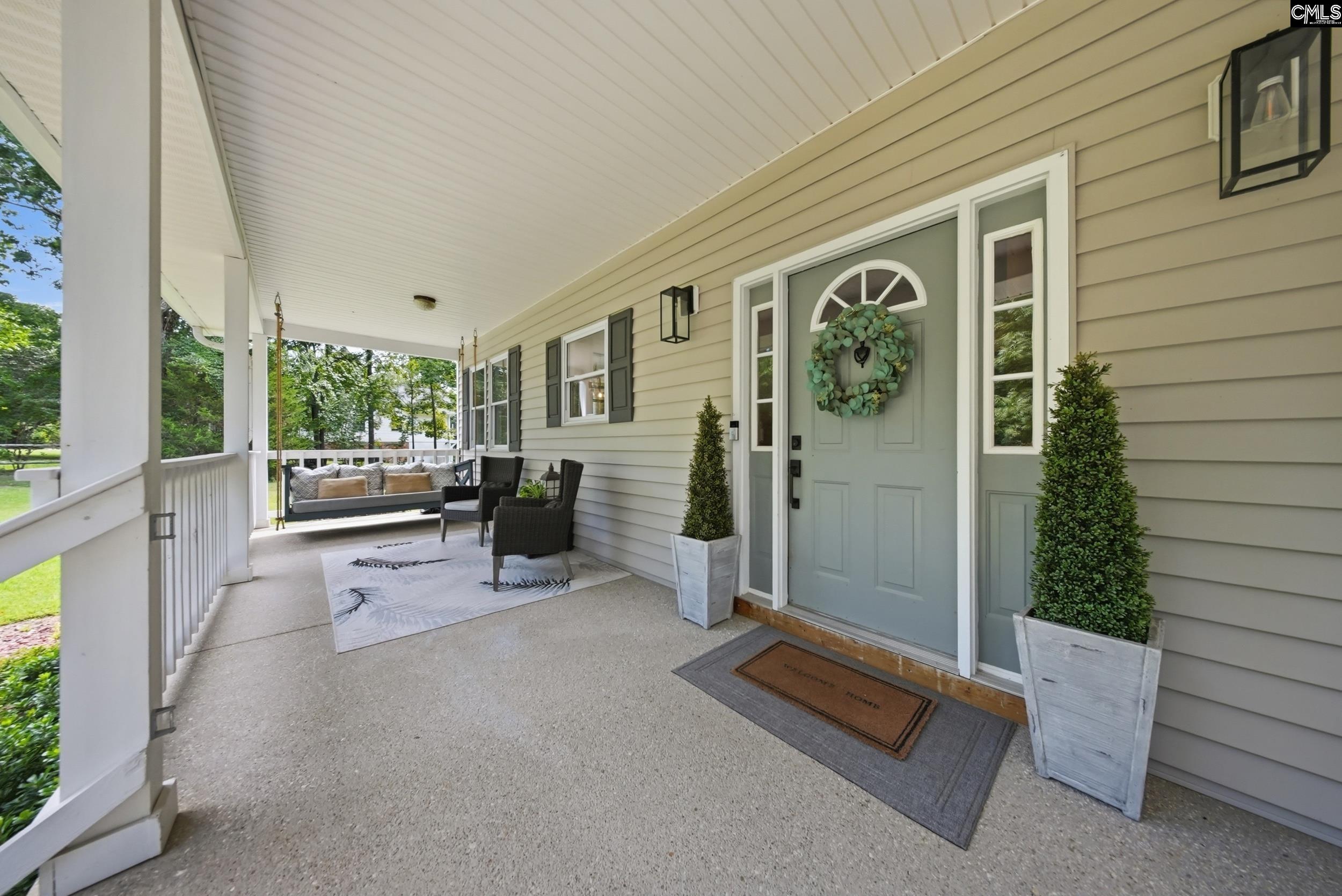 Covered front porch with a sage door, wreath, potted shrubs, and seating area.