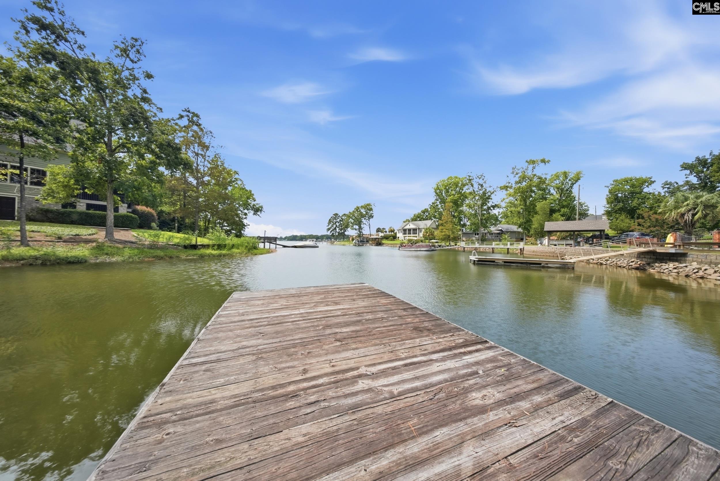Wooden dock extends into calm water, with tree-lined shores, waterfront homes, and boats.