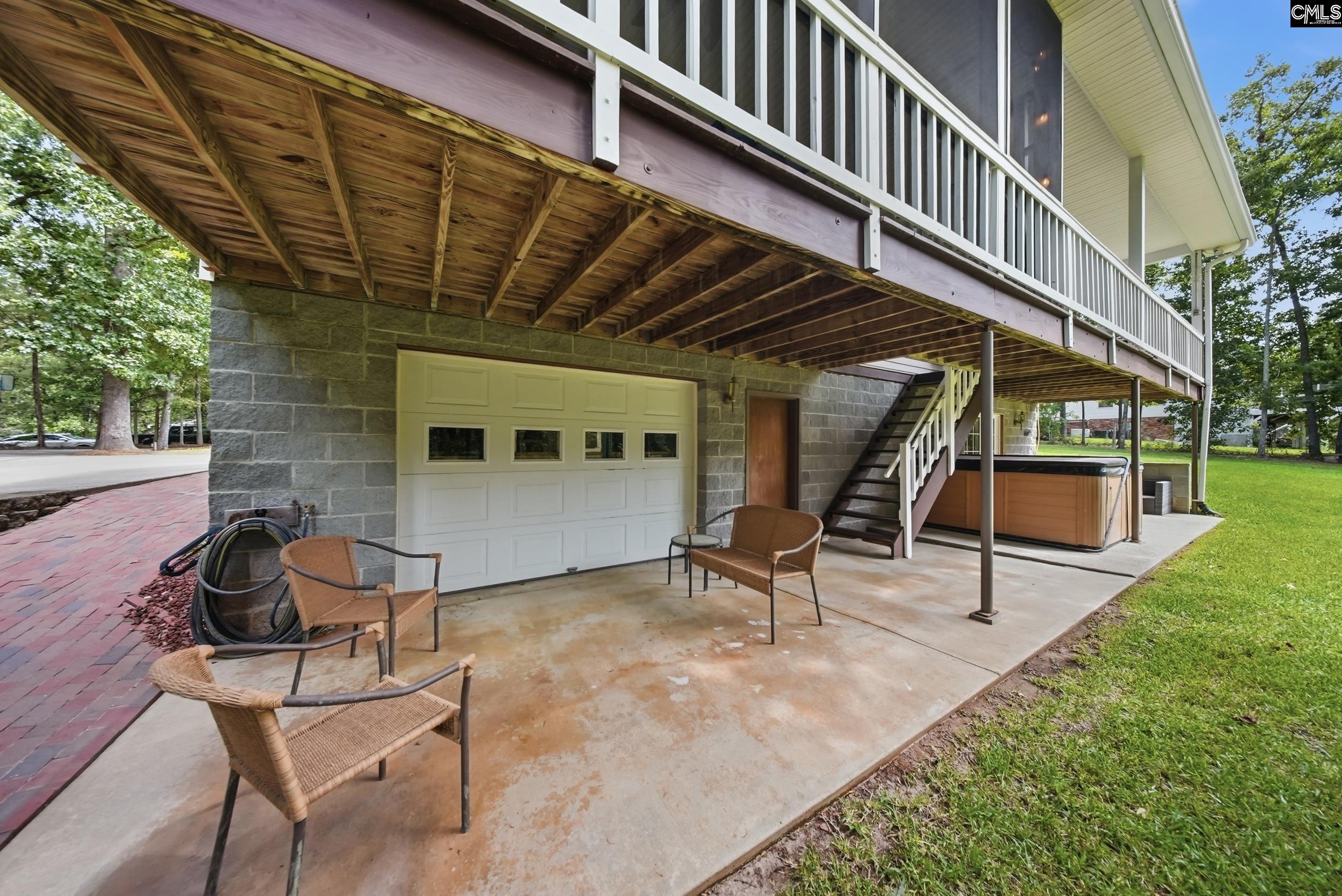 Covered patio beneath a raised wooden deck, with wicker chairs, exterior stairs, and a hot tub.