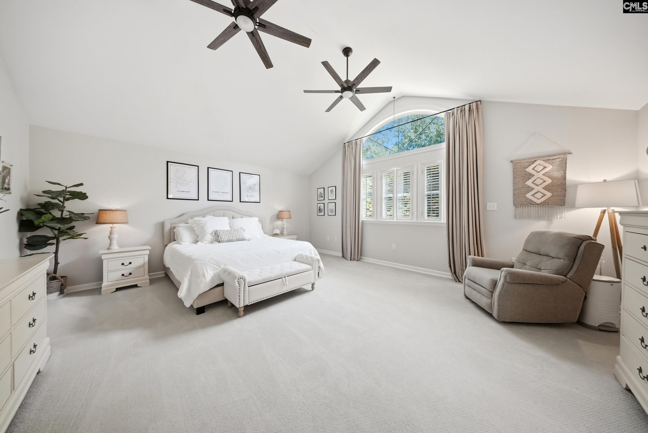 Furnished bedroom with a white bed, vaulted ceiling, and tall arched window.