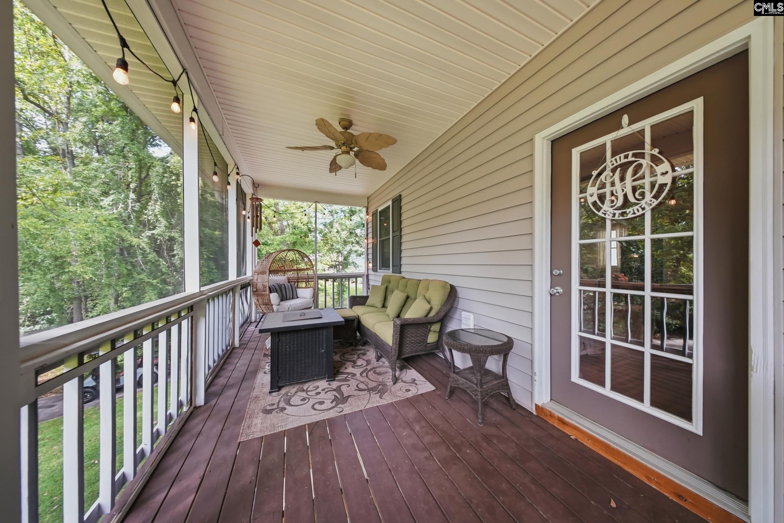 Covered porch with wicker seating, green cushions, string lights, and leafy trees beyond the railing.
