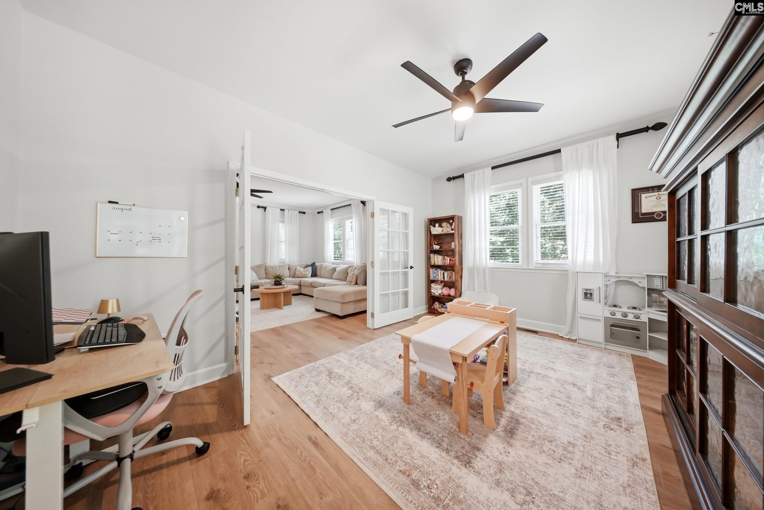 Bright home office and playroom with a desk, child-sized table, large curtained windows, and French doors to the living room.