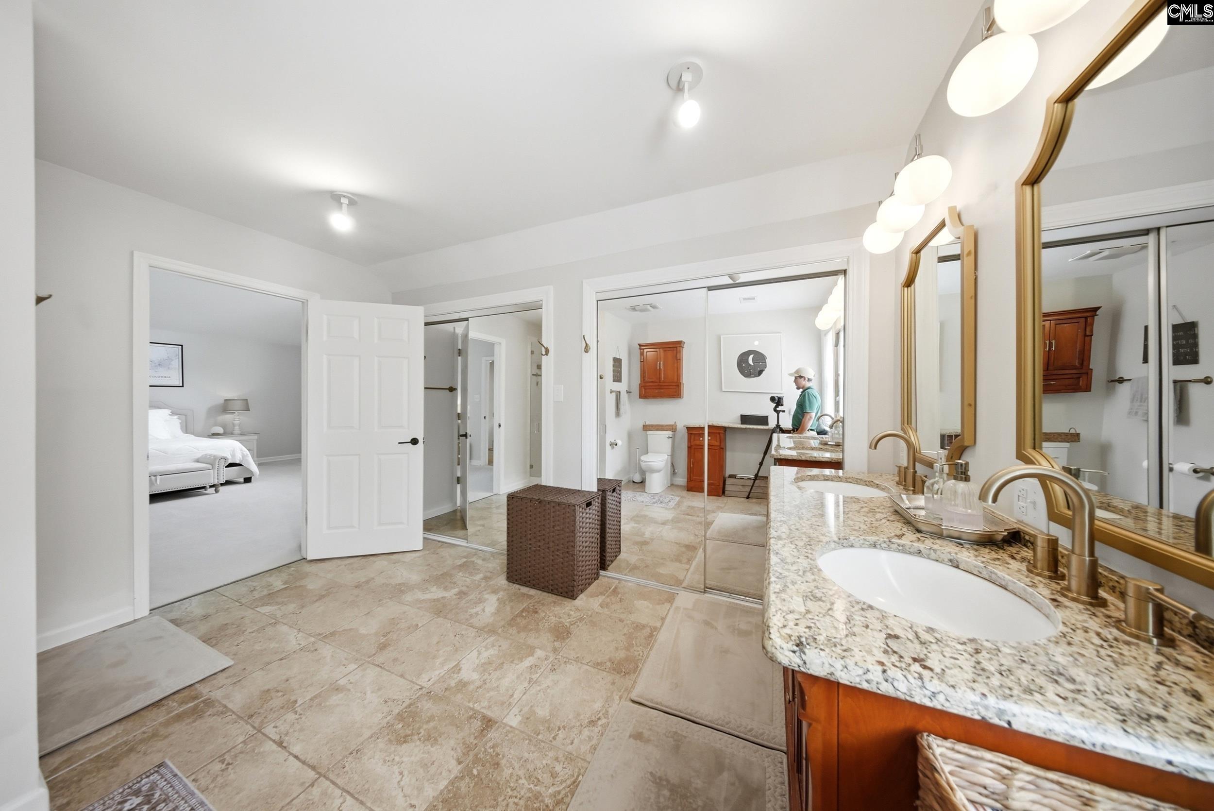 Bathroom with granite double vanity, gold-framed mirrors, beige tile floors, and glass shower enclosure.