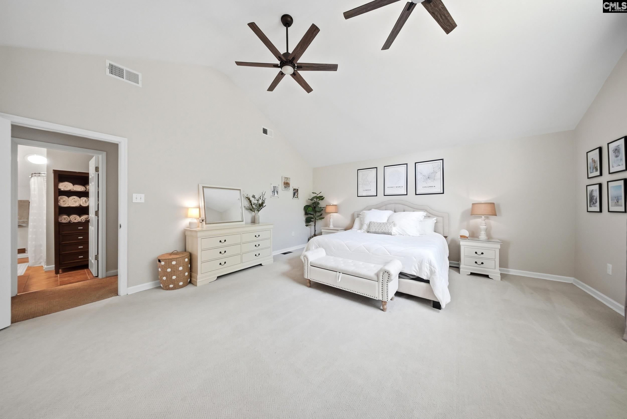 Vaulted primary bedroom with a white bed, upholstered bench, cream dresser, and adjoining bathroom doorway.