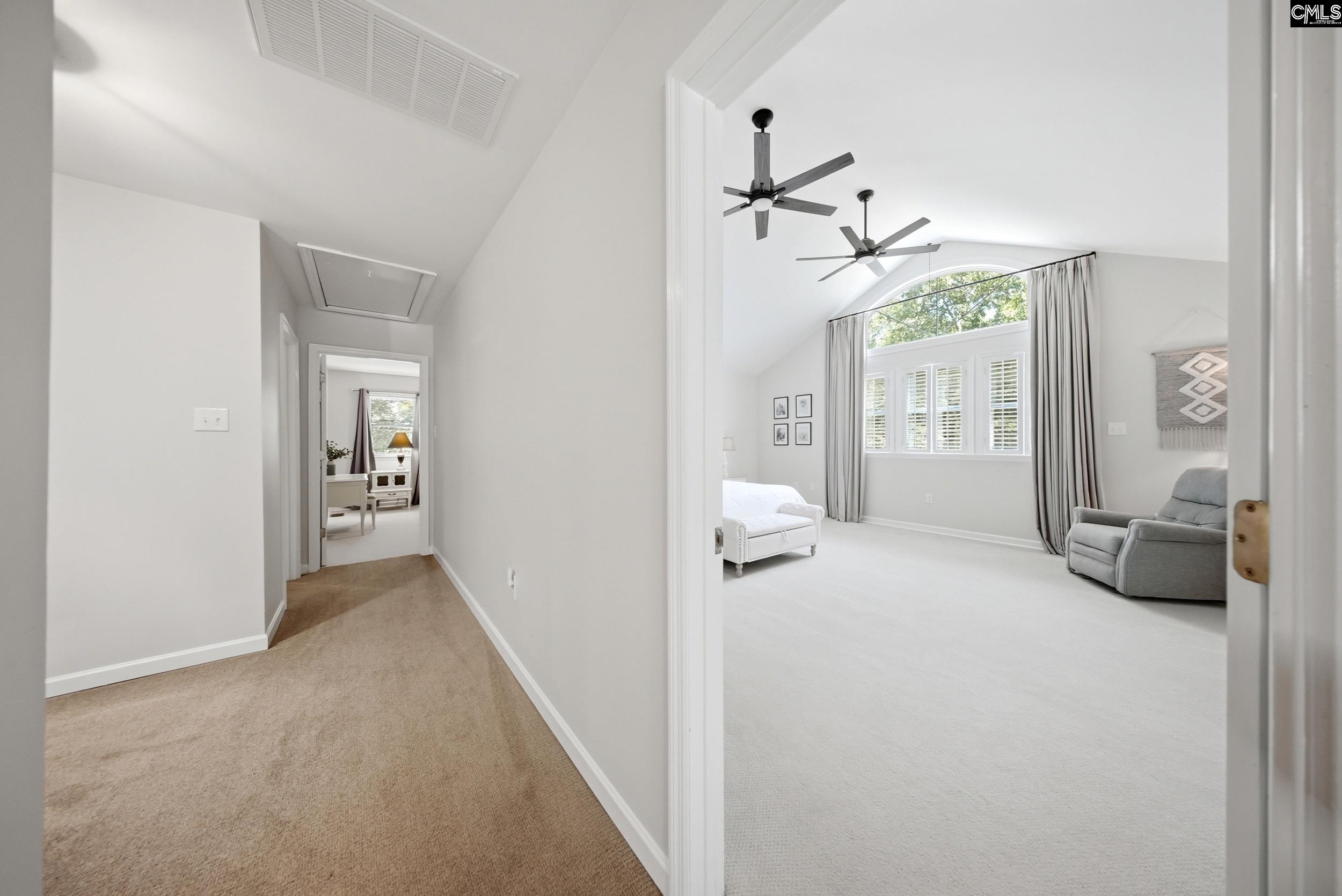 Bedroom with vaulted ceiling, arched window, ceiling fans, and gray seating, viewed from carpeted hallway.
