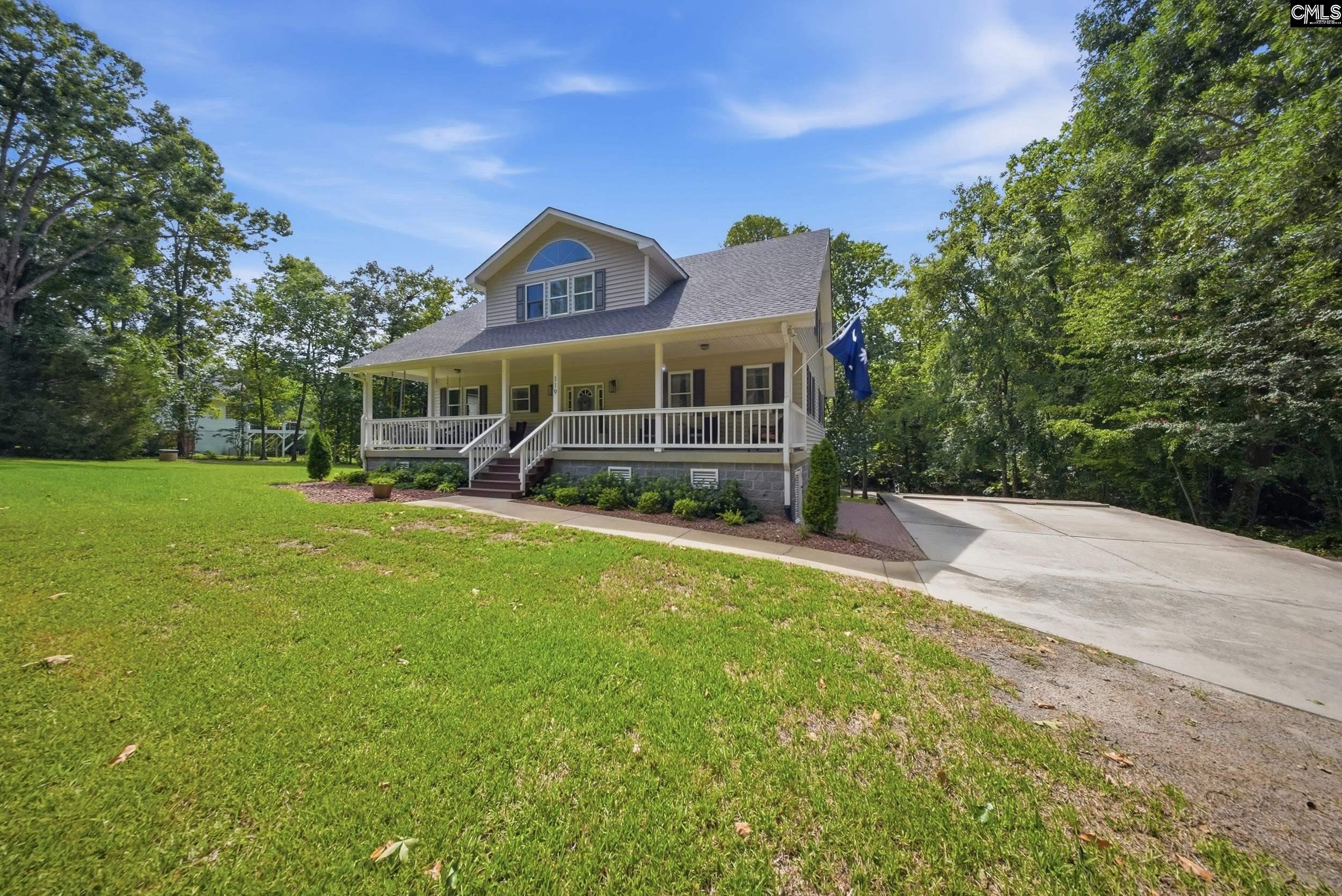 Beige two-story home with a covered porch, broad lawn, and wide driveway among mature trees.