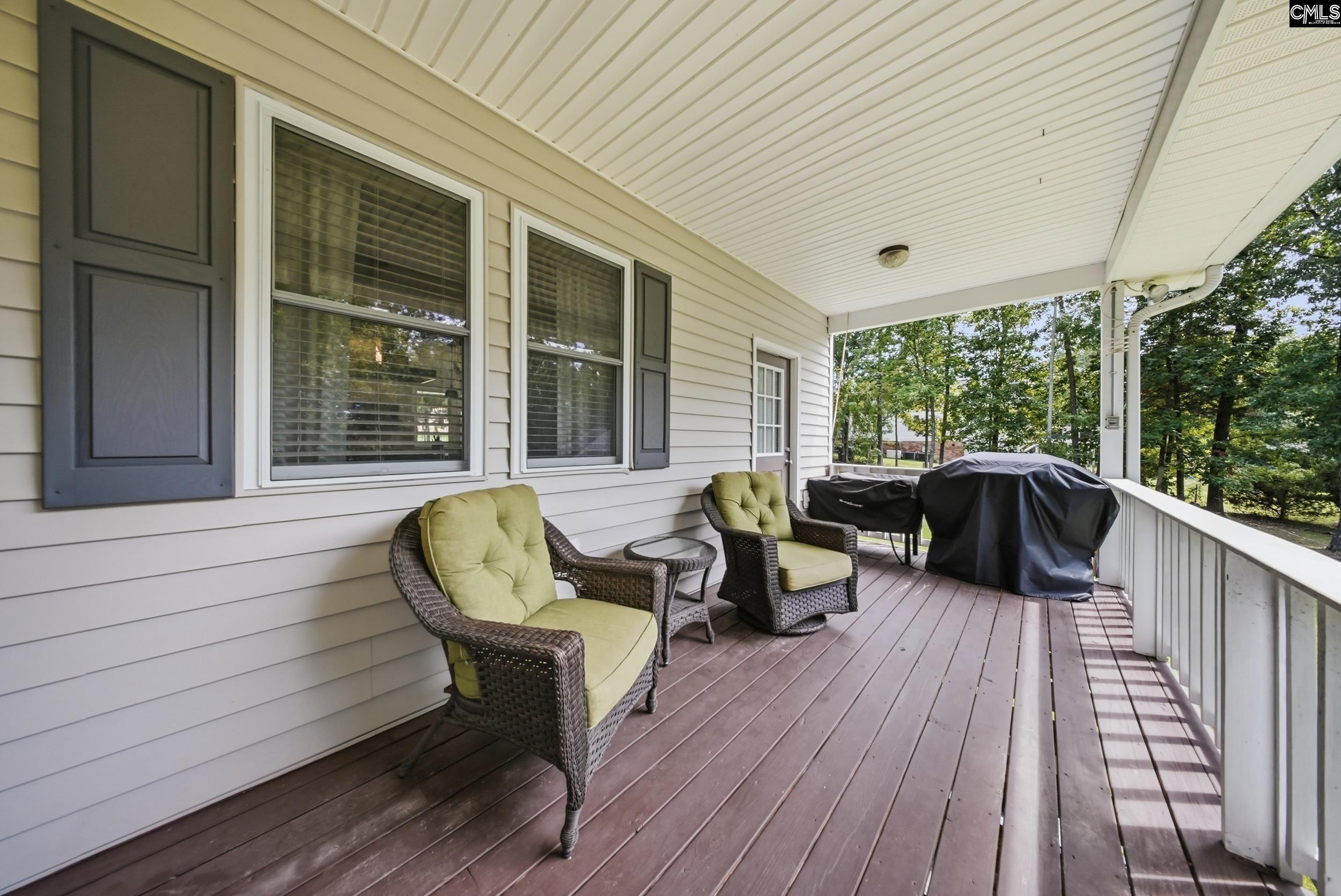 Covered porch with wicker chairs, green cushions, and covered grills beside a wooded yard.