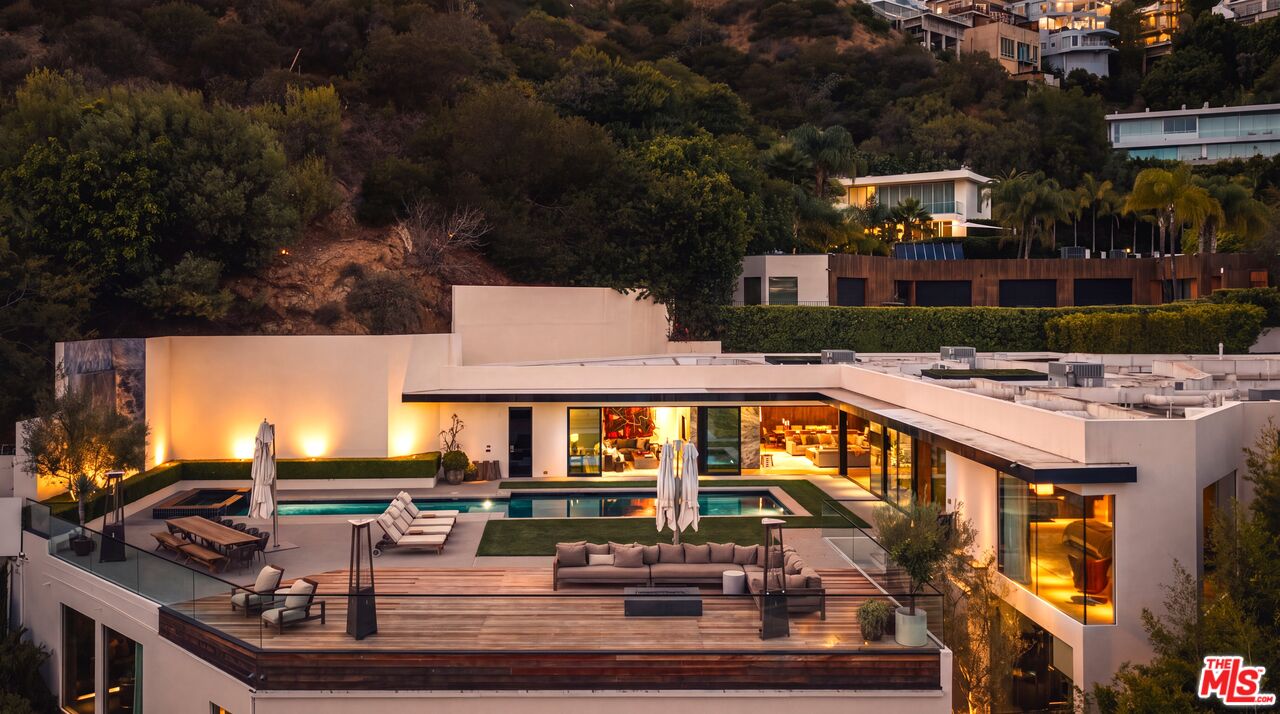 Modern hillside home with rooftop pool and furnished deck, illuminated at dusk.
