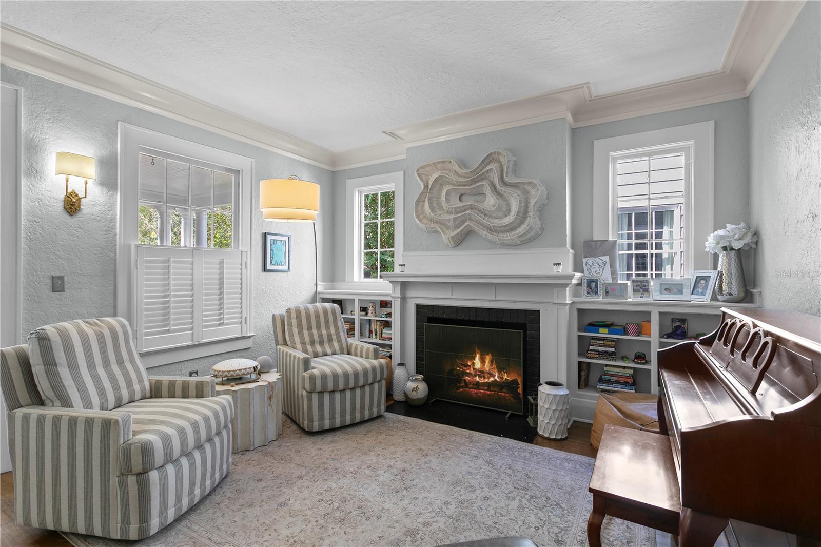 Living room with a lit fireplace, built-in shelves, striped armchairs, and a wooden upright piano.