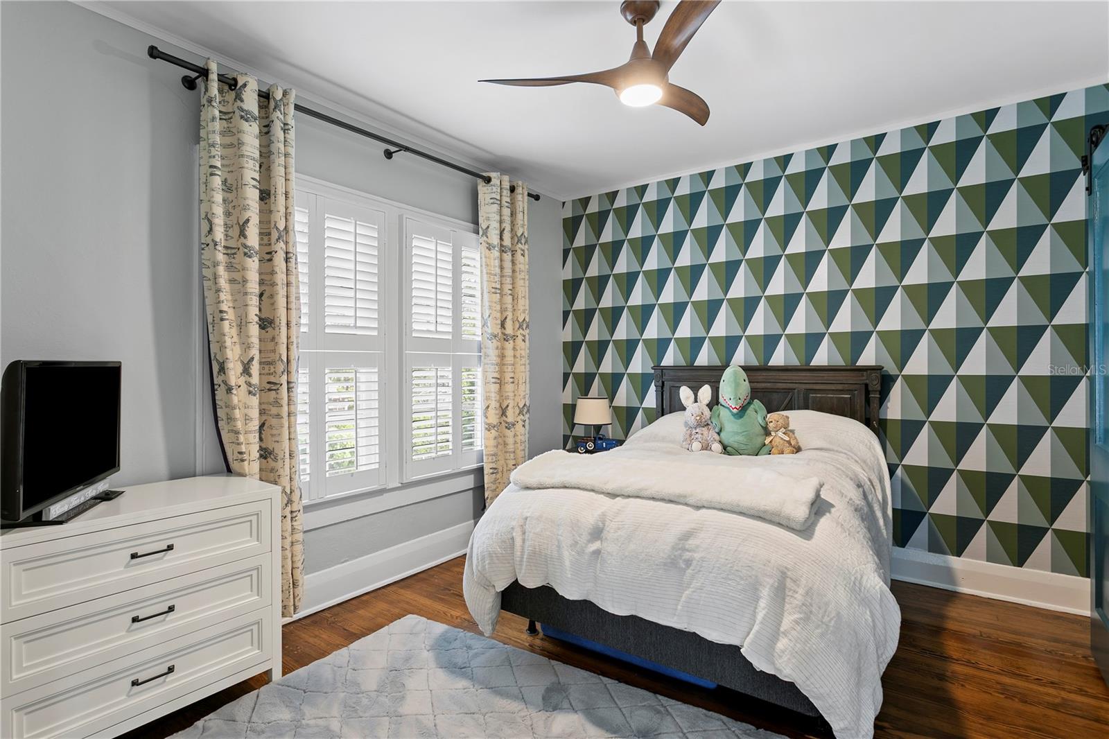 Bedroom with geometric accent wall, shuttered windows, and a bed topped with stuffed animals.