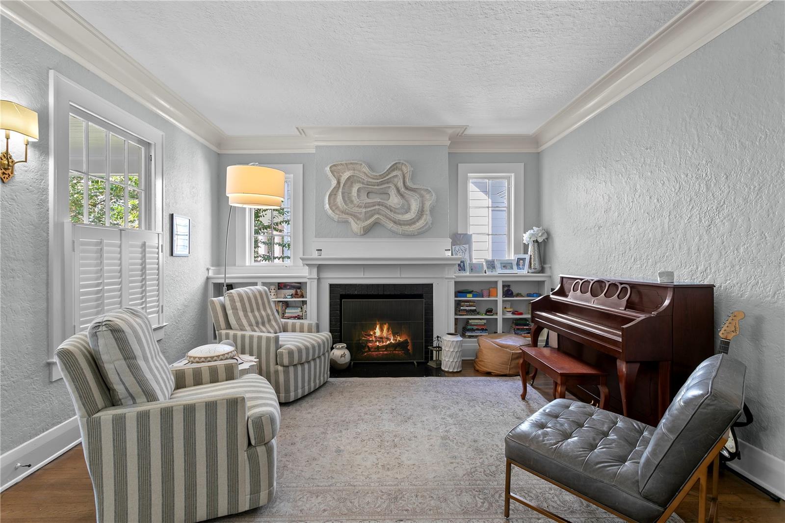 Living room with a lit fireplace, striped armchairs, built-in shelves, and an upright piano.