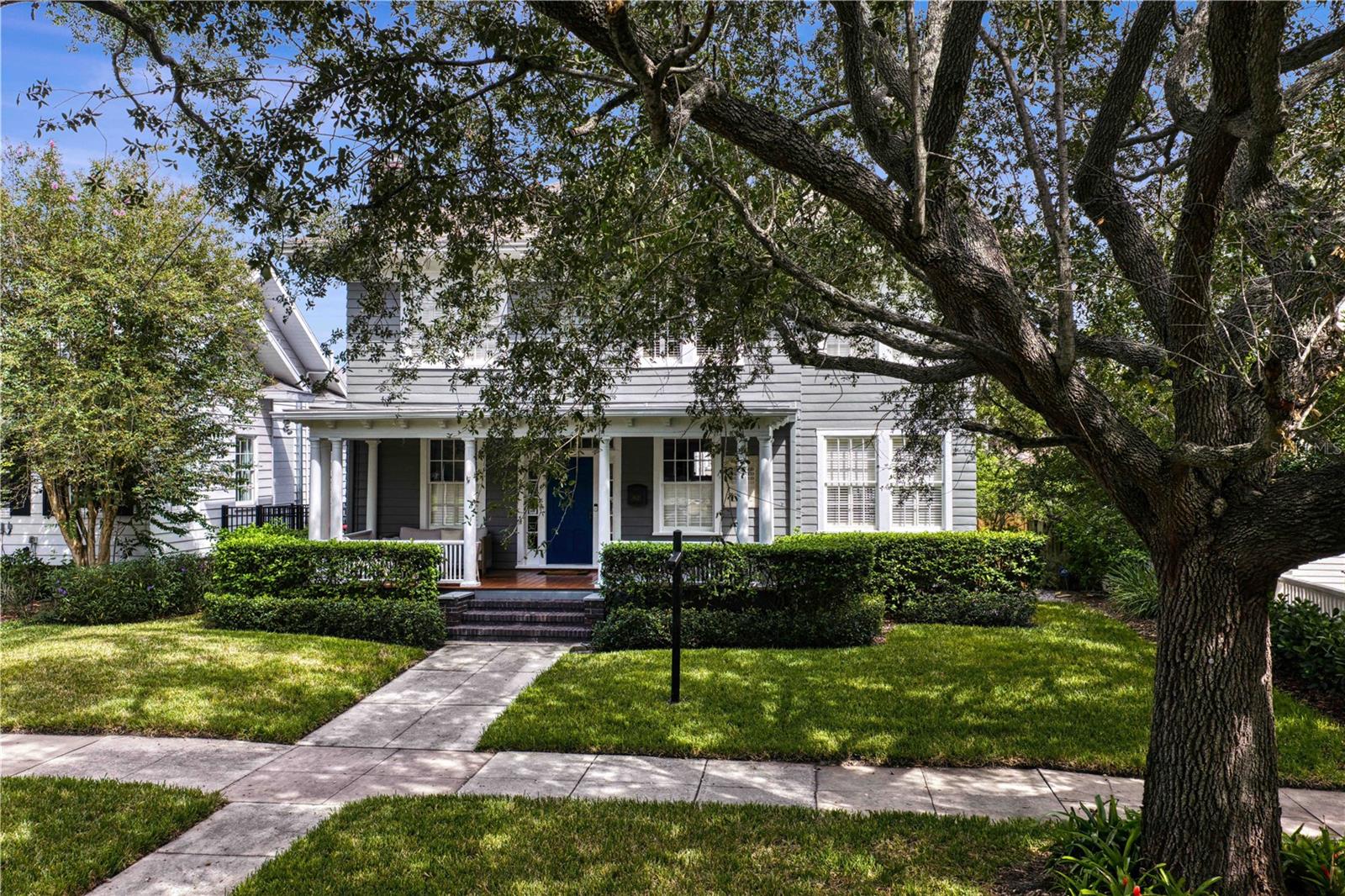 White clapboard house with a covered porch and blue front door beneath a mature oak tree.