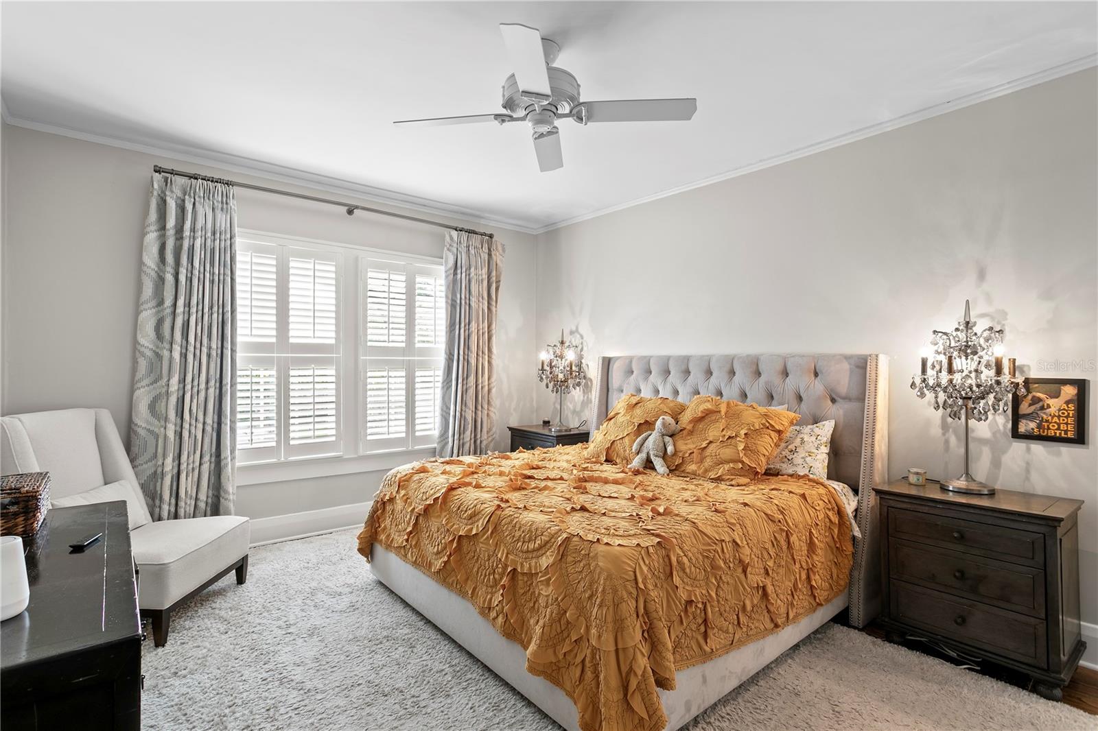 Furnished bedroom with a mustard-gold ruffled bedspread, tufted gray headboard, and bright shuttered windows.