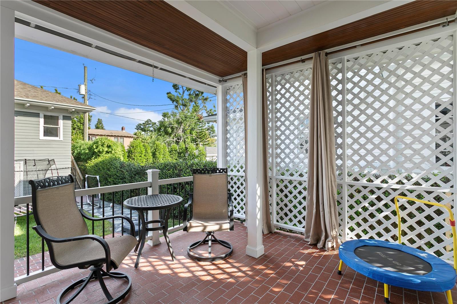 Covered patio with brick flooring, lattice screens, outdoor seating, and a small trampoline.
