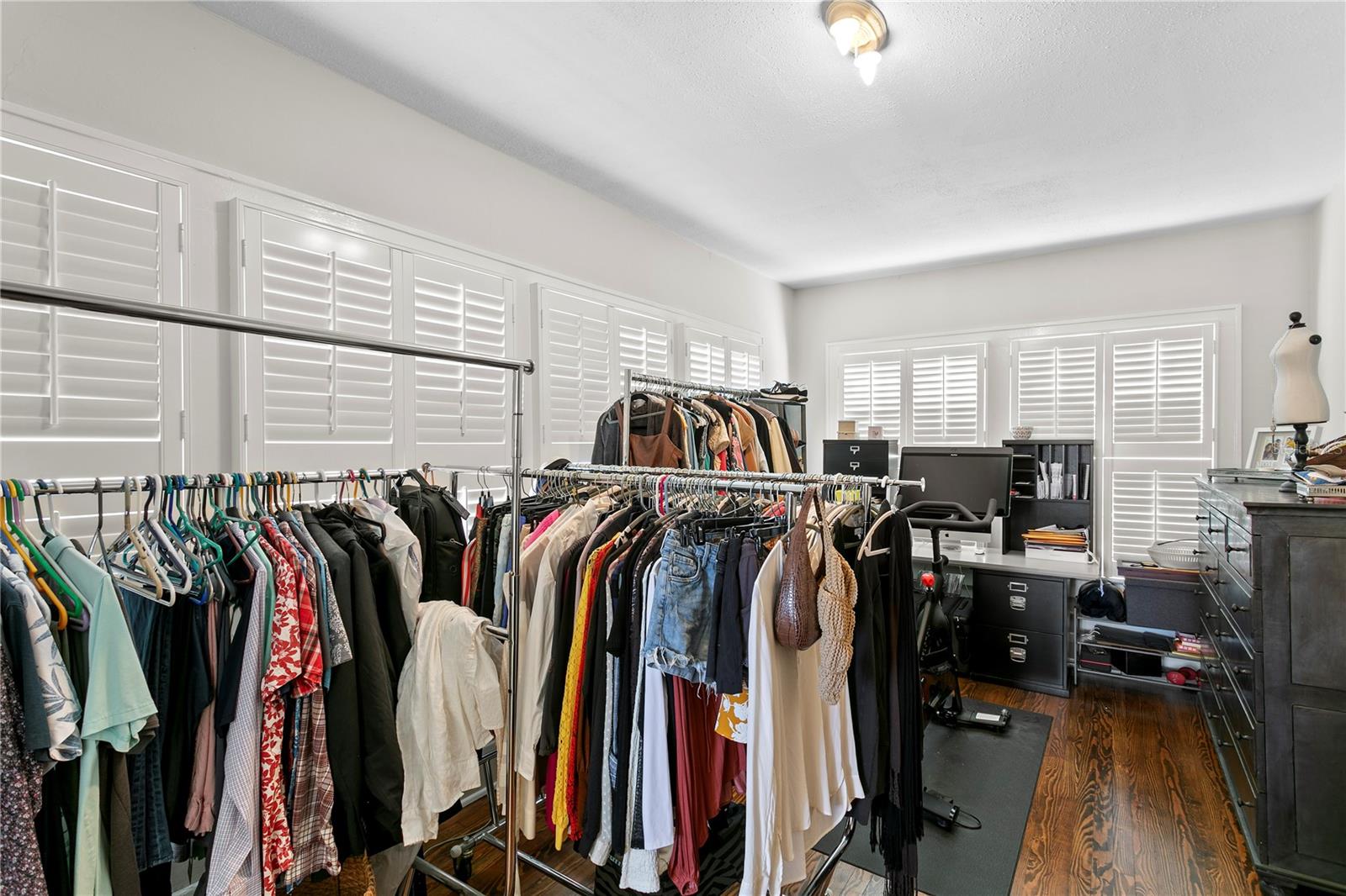Walk-in closet with clothing racks, plantation shutters, dark wood floors, and a desk.
