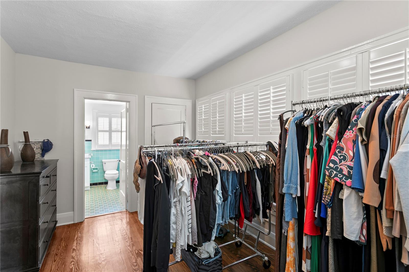 Walk-in closet with clothing racks, plantation shutters, and a doorway to a turquoise-tiled bathroom.