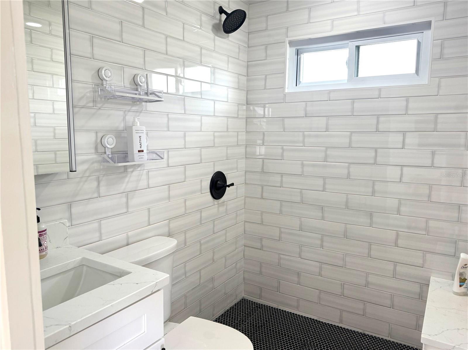 Bathroom shower with glossy white subway tile, black fixtures, and a small high window.