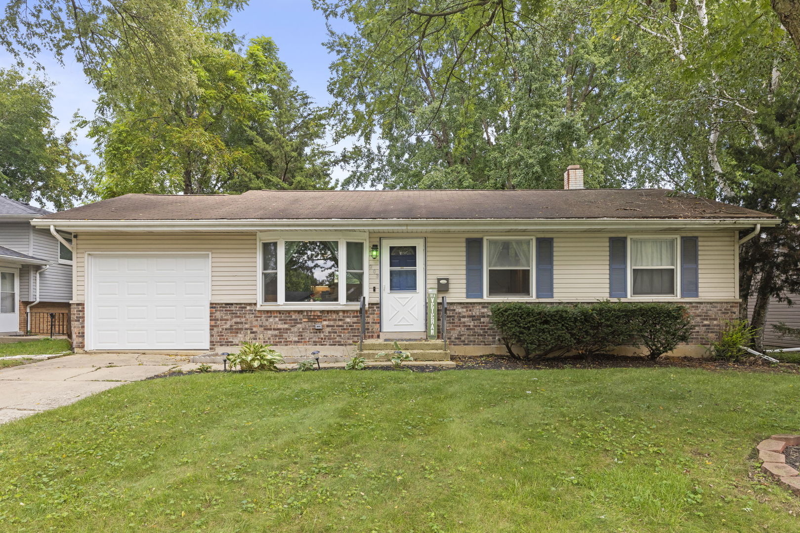 Single-story ranch house with beige siding, brick facade, attached garage, and a green lawn.