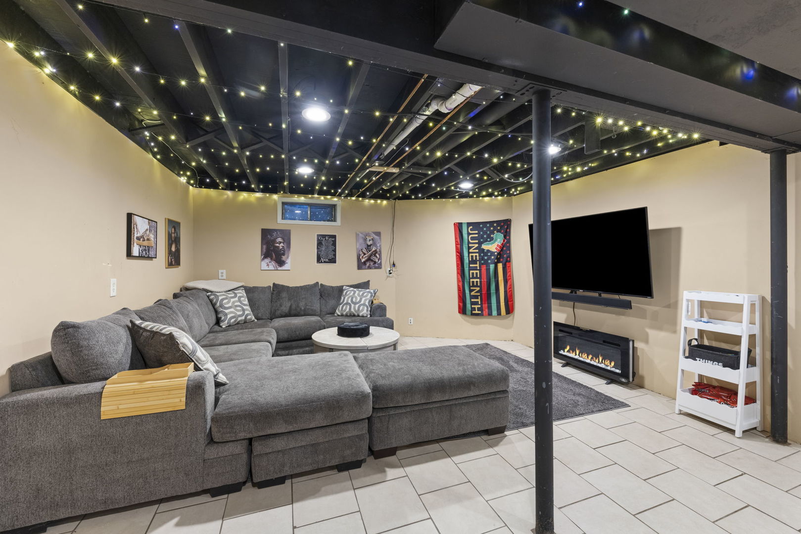 Basement living room with a gray sectional, string-lit exposed ceiling, television, and electric fireplace.