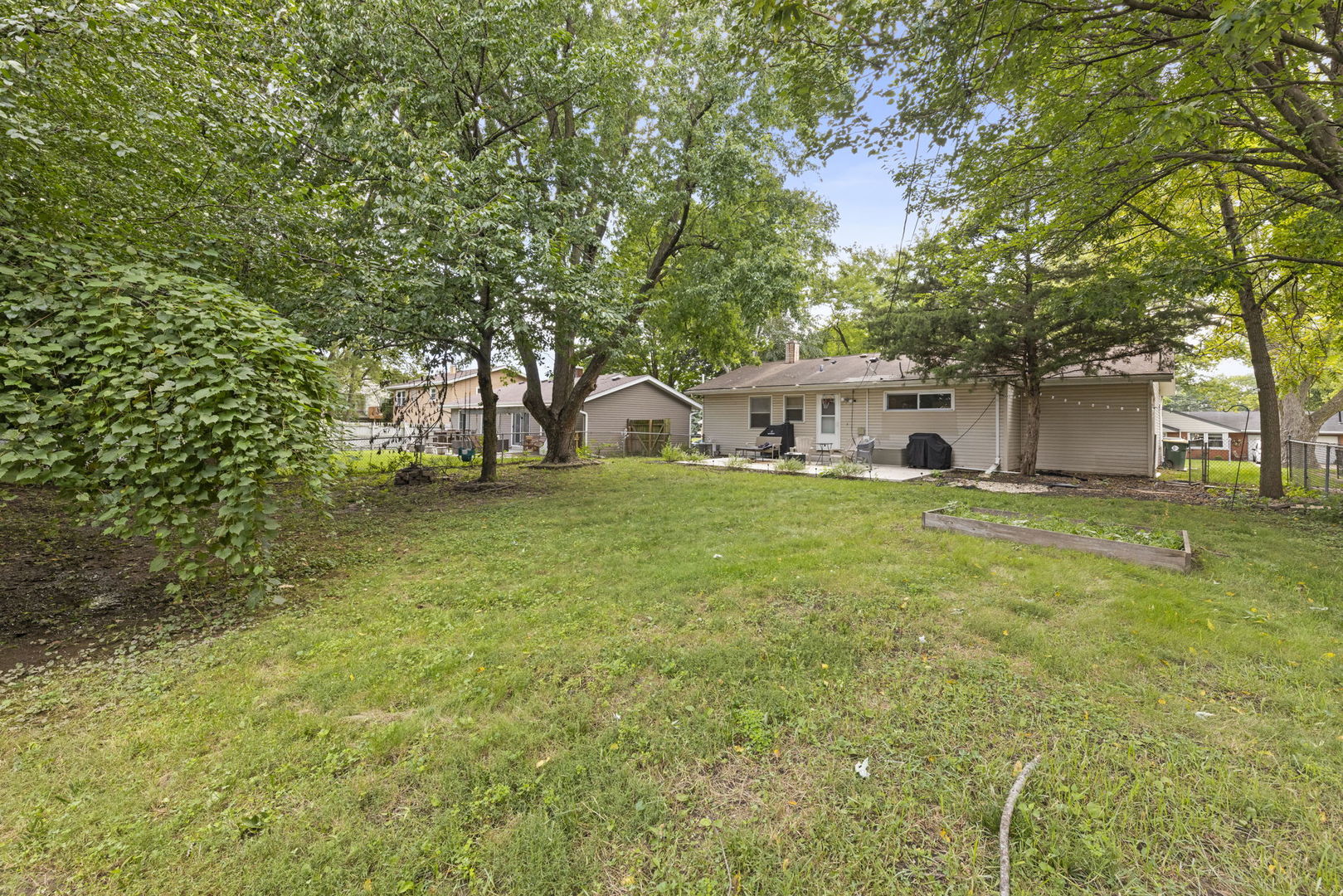 Back of a beige single-story home with a tree-shaded lawn, patio, and raised garden bed.