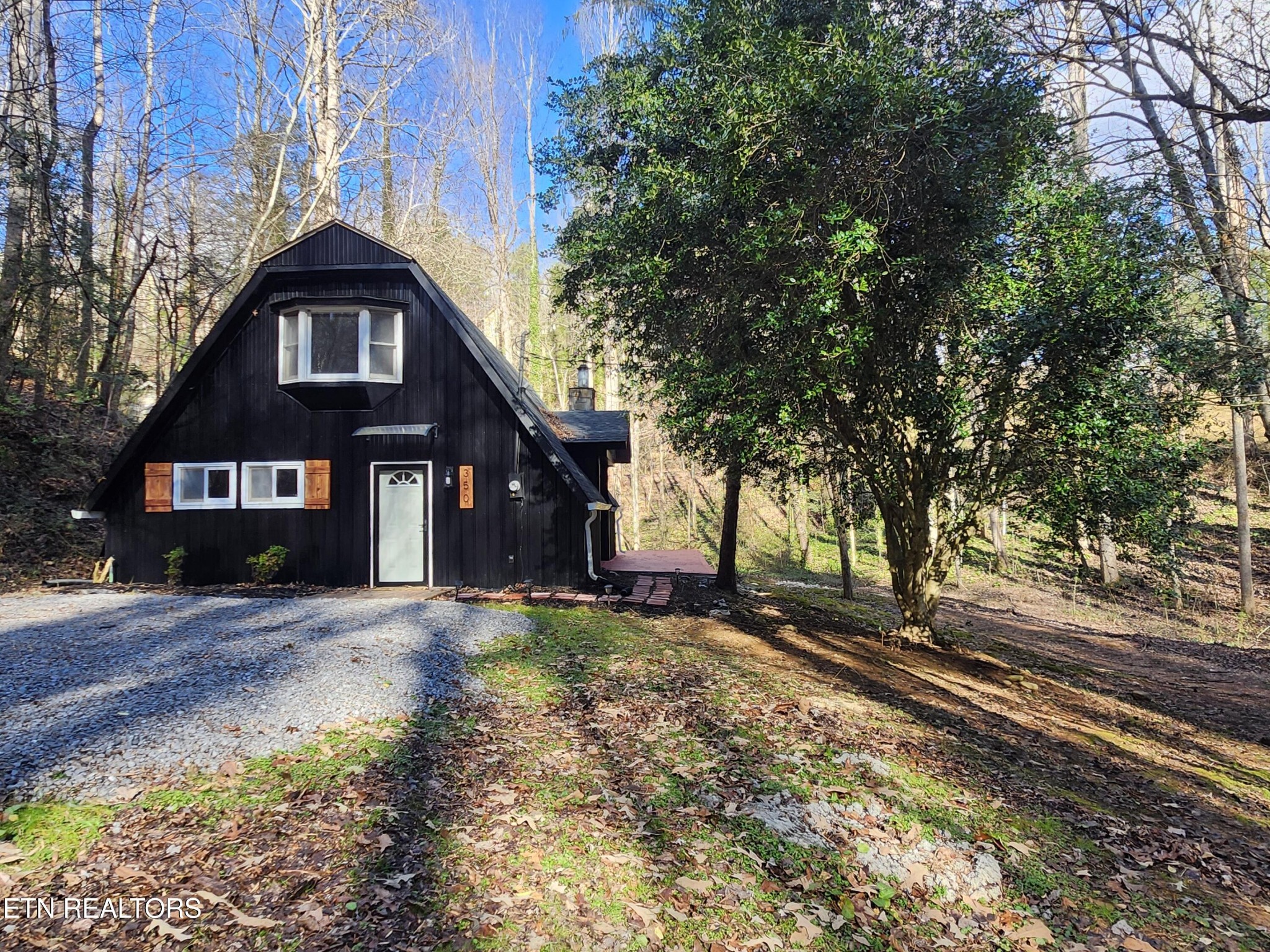 Black steep-roofed cabin with white-trimmed windows beside a gravel driveway in a wooded setting.