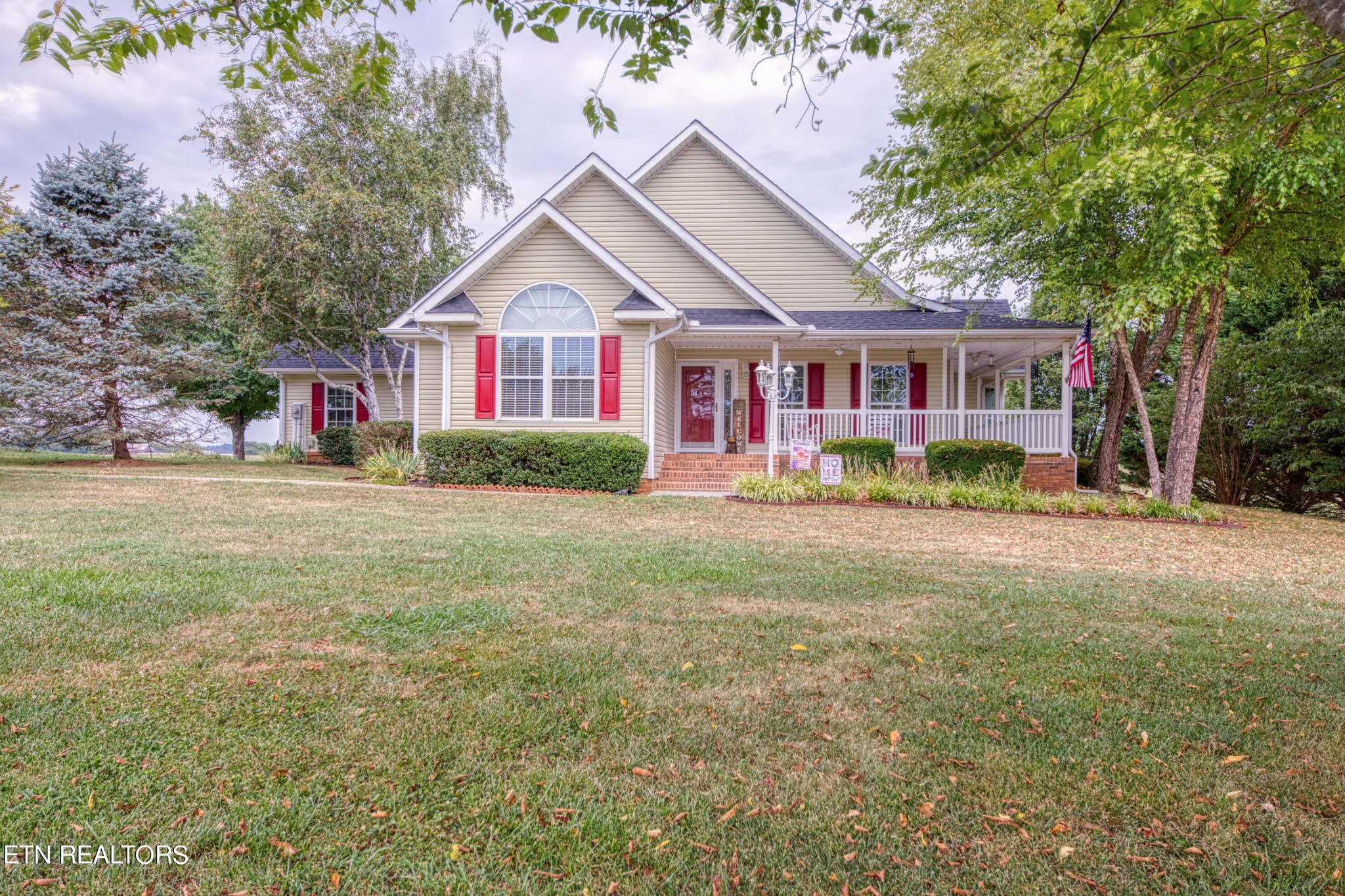 Beige gabled house with red shutters, a covered porch, and a broad front lawn.