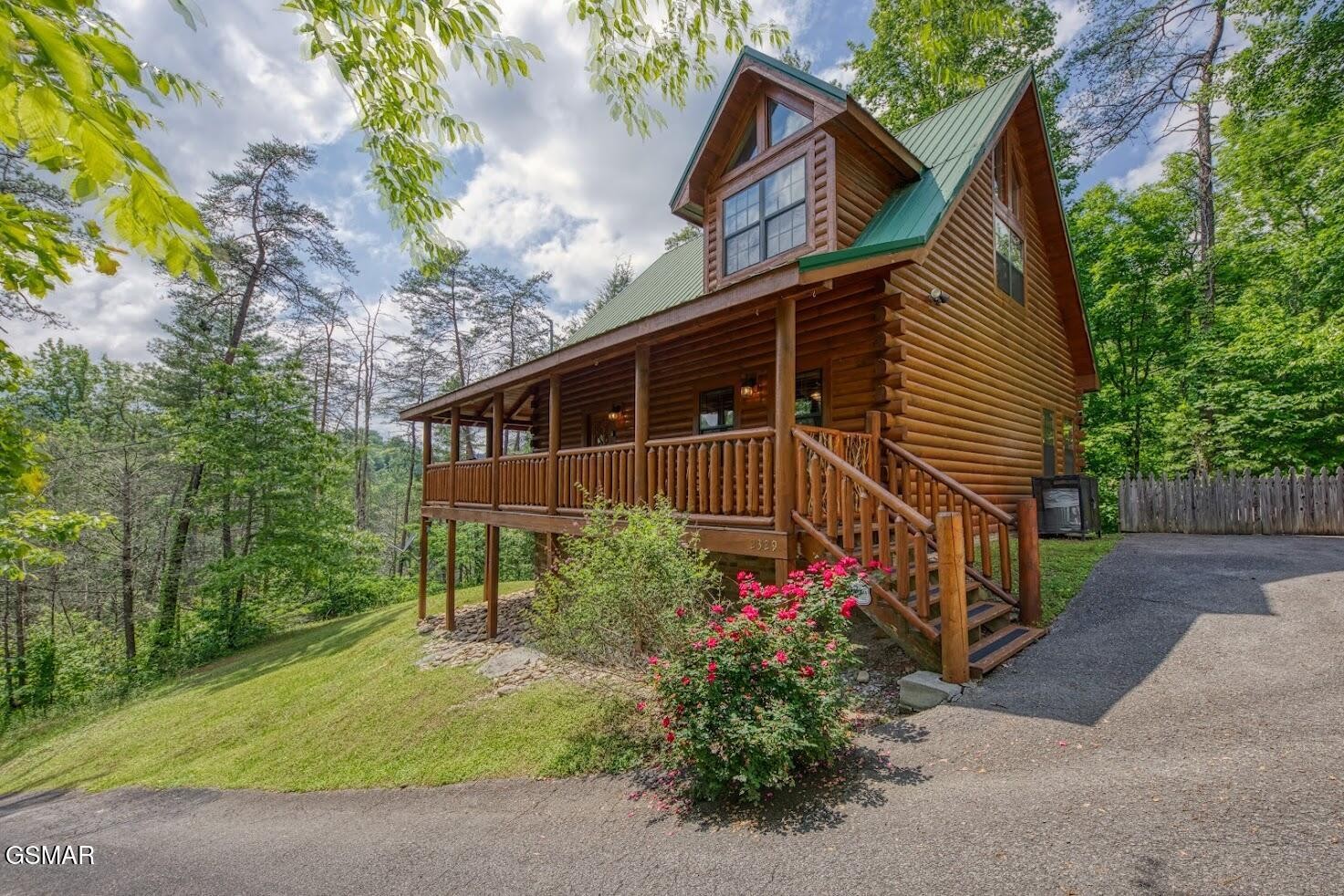 Two-story log cabin with a green metal roof, covered porch, and pink flowers among trees.