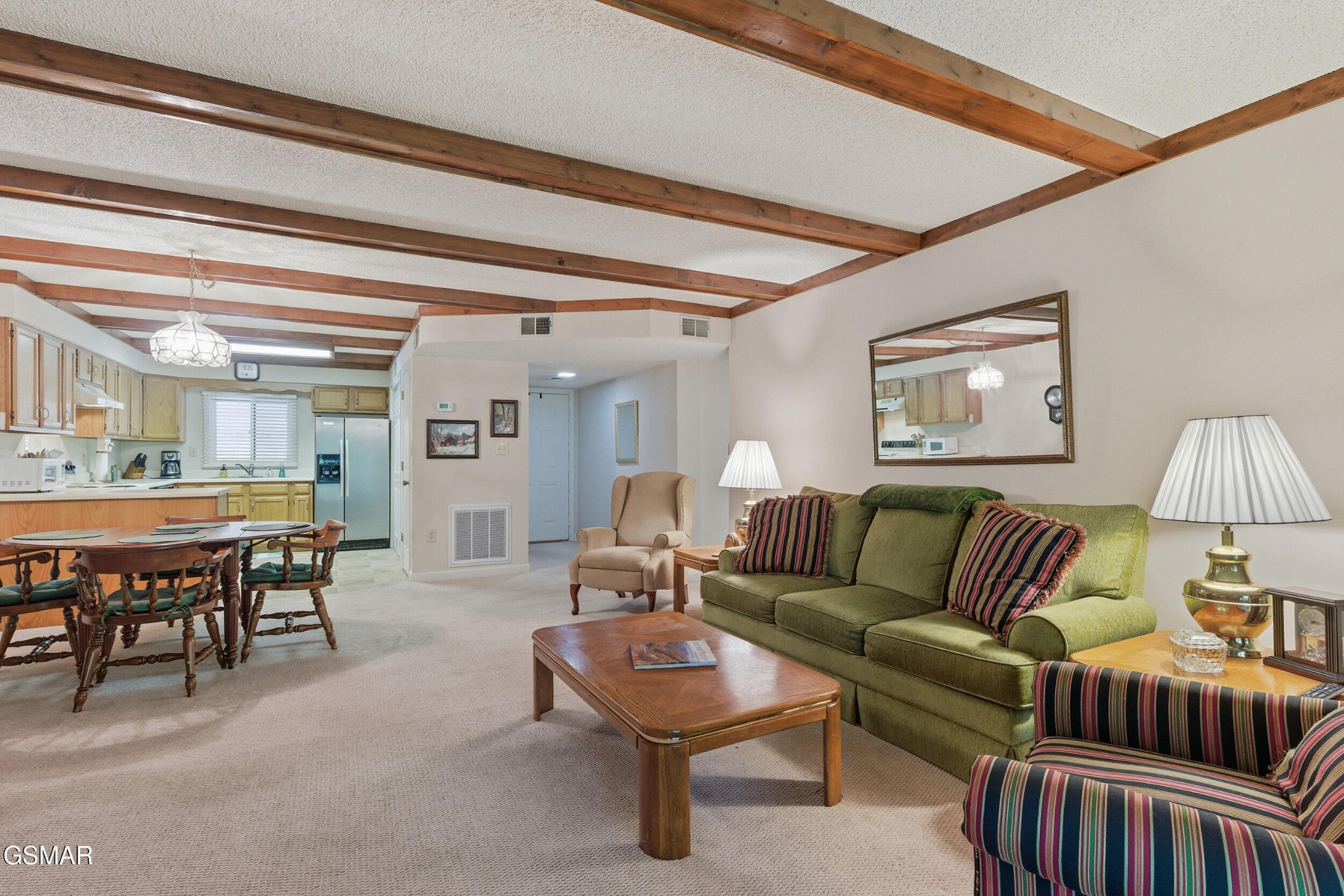 Open-plan living room with green sofa, striped armchairs, exposed wood ceiling beams, and adjoining kitchen.