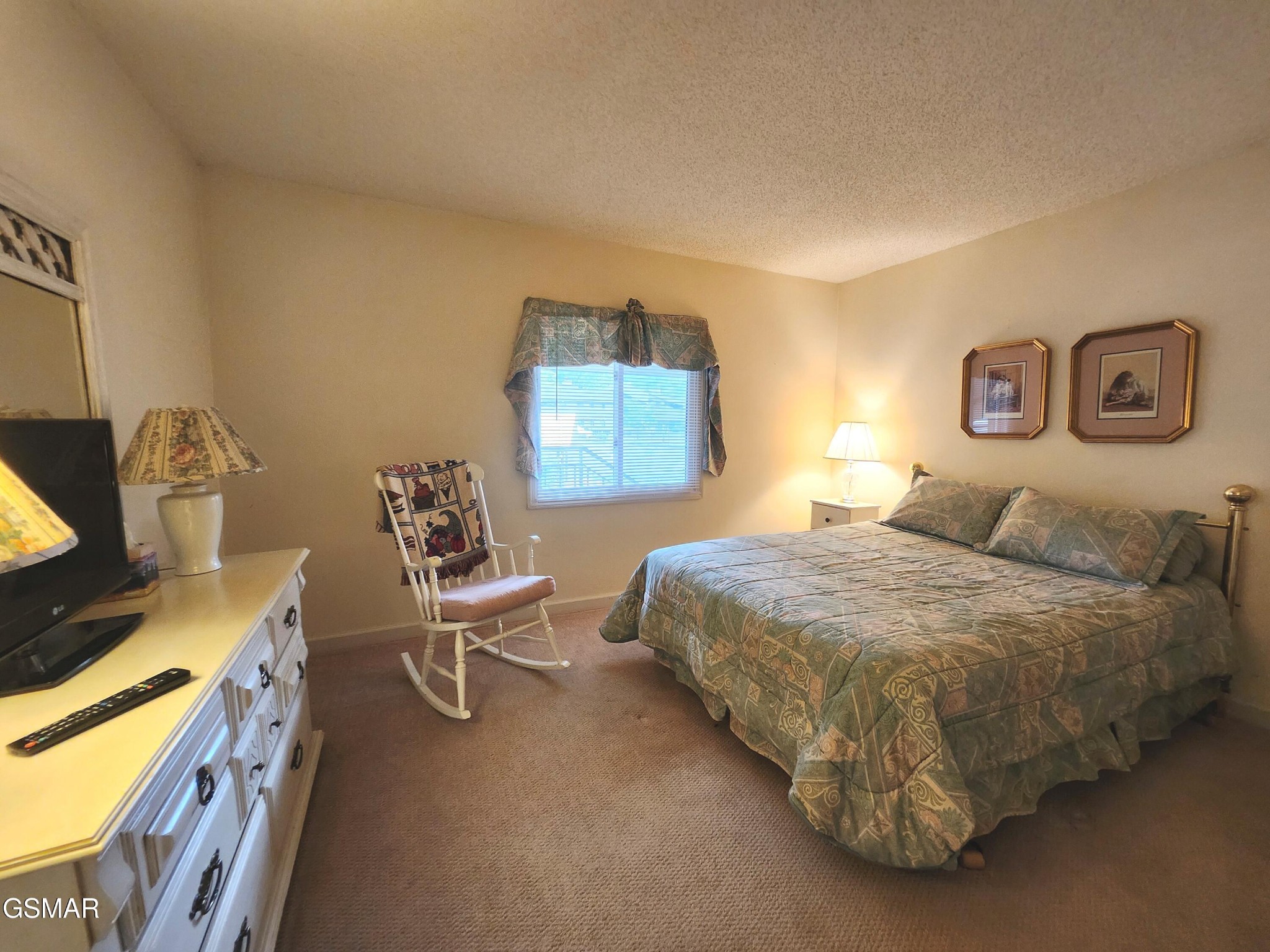 Furnished bedroom with a patterned bedspread, rocking chair, and window with blinds.