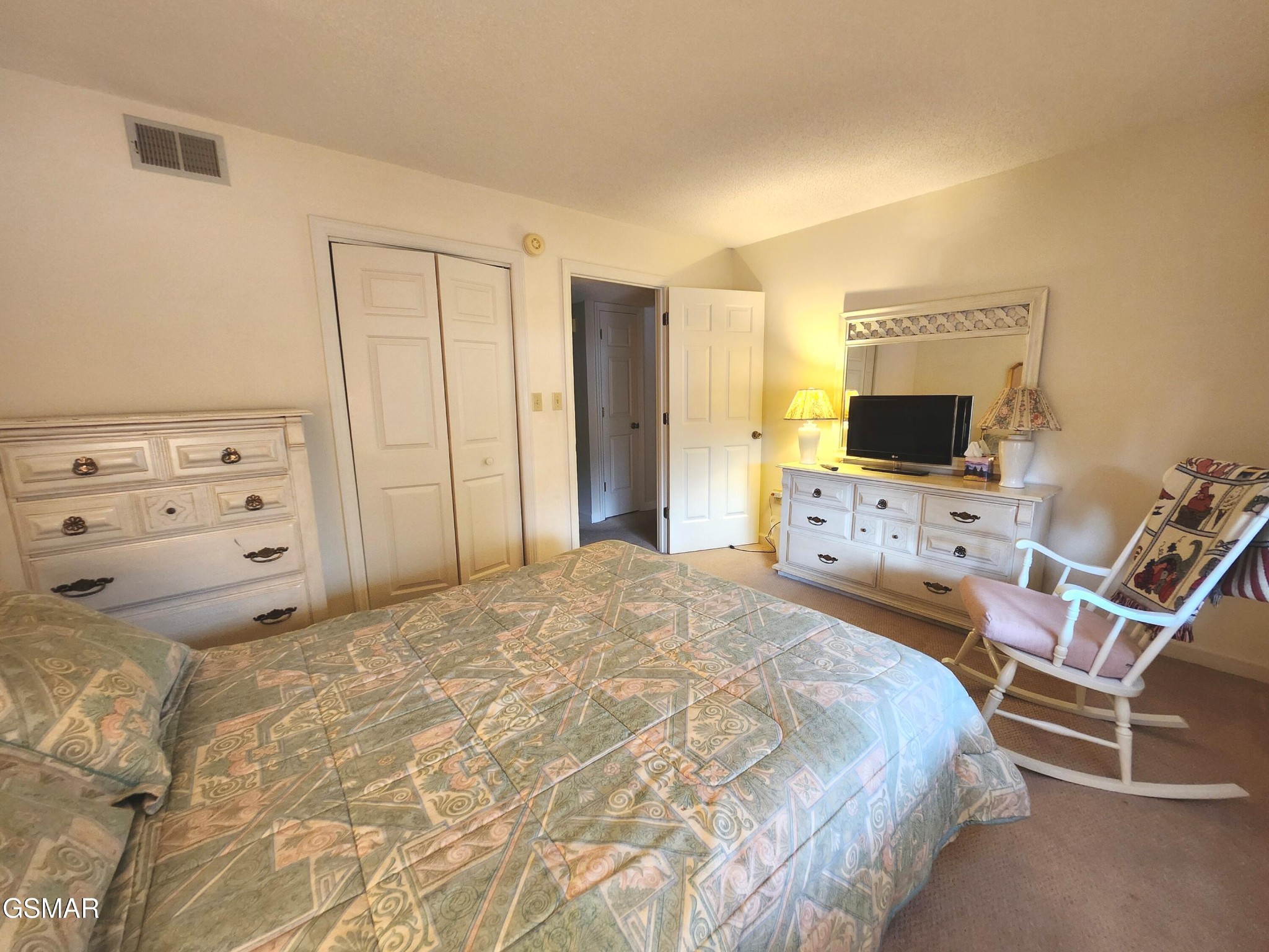 Furnished bedroom with a patterned quilt, white dressers, television, and cushioned rocking chair.